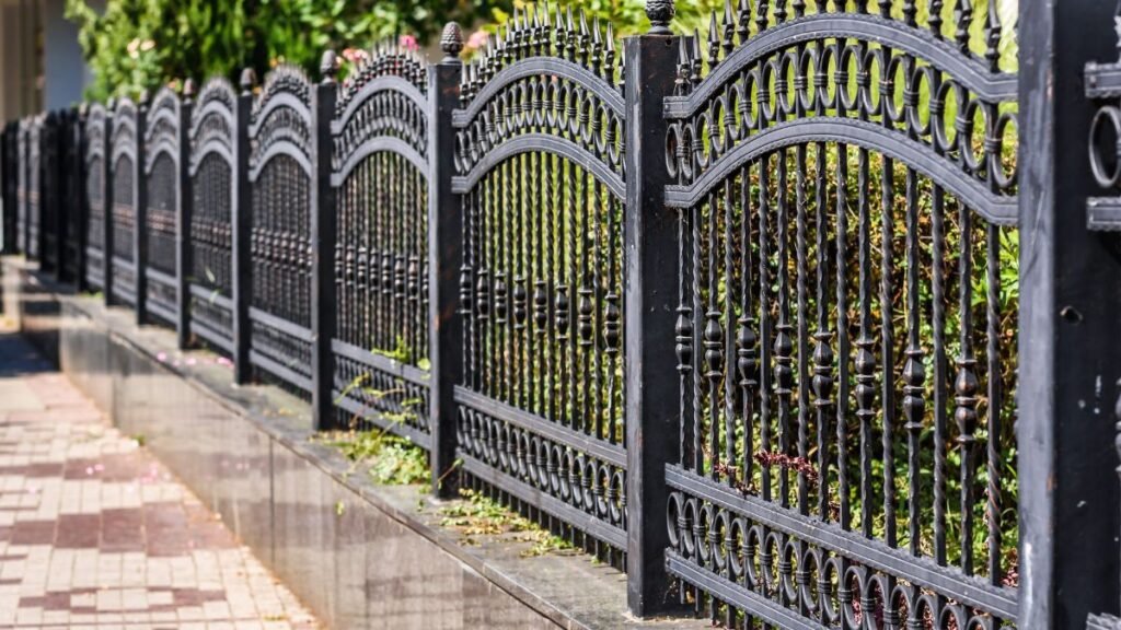 Fekete vas kerítés, amely egy rendezett kertet határol, zöld növényekkel.