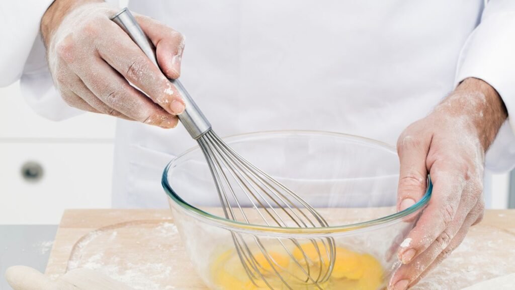 Egy kéz habverőt használ, tojásokat kever egy tálban, lisztporral körülvéve.