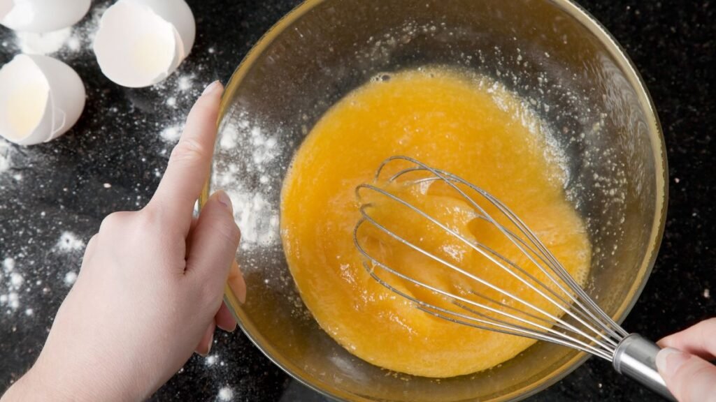 Egy kéz habverőt használ, hogy tojásos masszát keverjen egy tálban.