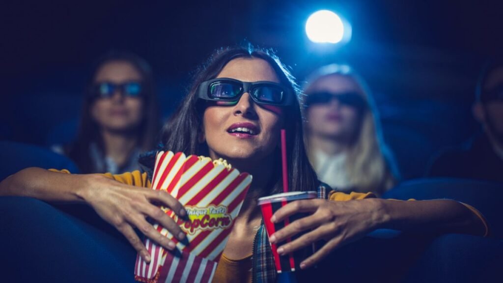 Egy nő popcornnal és üdítővel néz egy filmet a moziban, 3D szemüveget viselve.