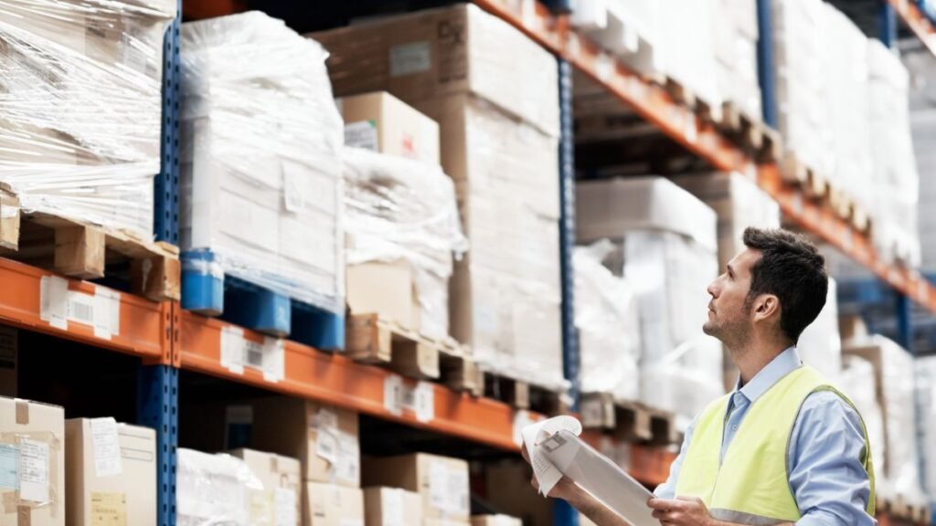 Egy férfi egy raktárban áll, dobozokat néz, miközben listát tart a kezében.