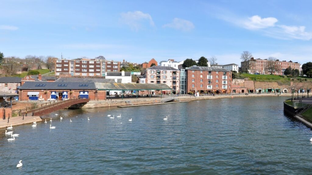 A rakparton sétáló emberek és úszó hattyúk a vízben, napfényes időben.