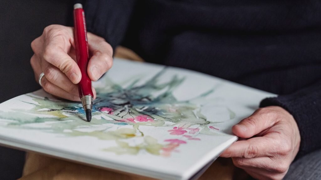 Egy kéz vízfestékkel rajzol egy virágos kompozíciót egy lapra.