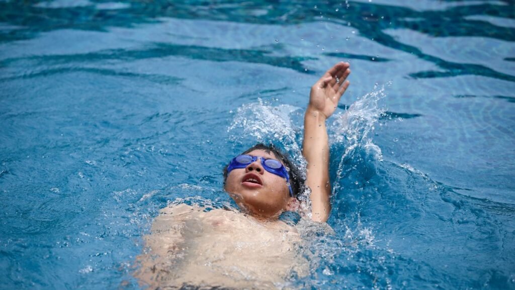 Egy fiú hátúszik a medencében, vízcseppek fröccsennek körülötte.