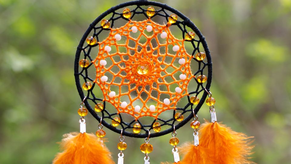 10 tipp, hogy könnyebben megértsd az álmaidat Egy színes álomfogó, narancssárga díszítéssel és tollakkal, zöld háttér előtt.