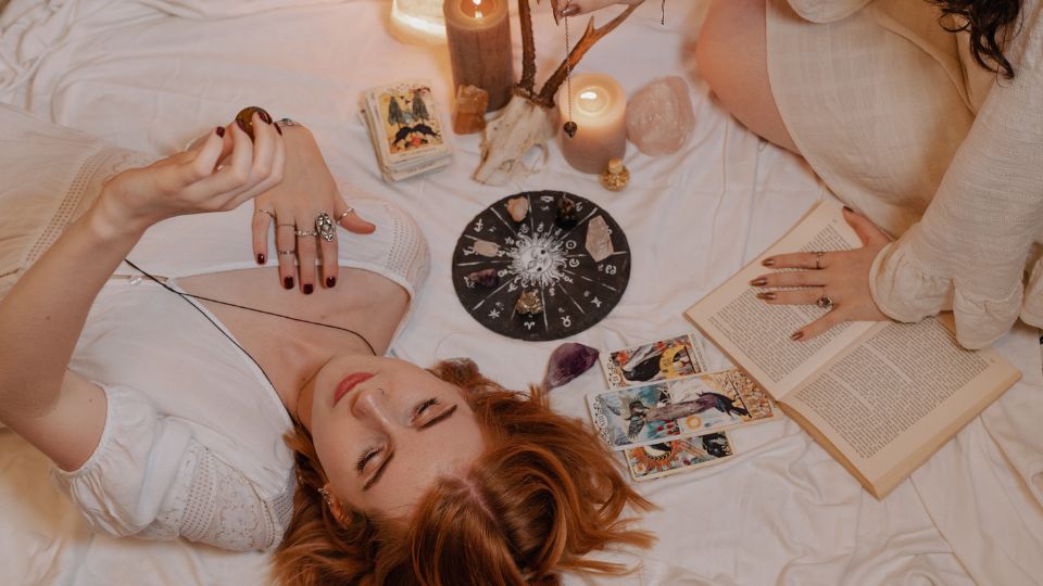 Két nő tarot kártyákat olvas, gyertyák és kristályok között ülve.