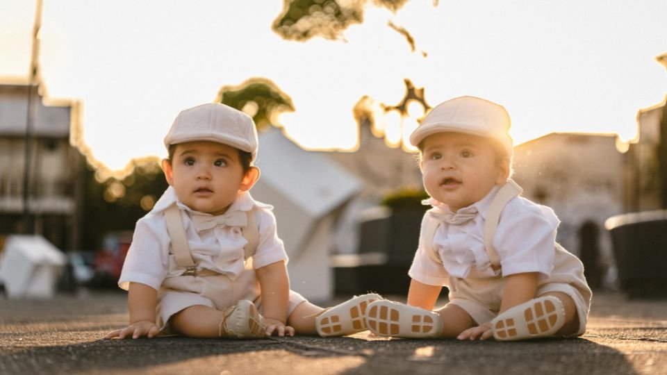 Két kisbaba ül a földön, elegáns ruhában, napfényben.