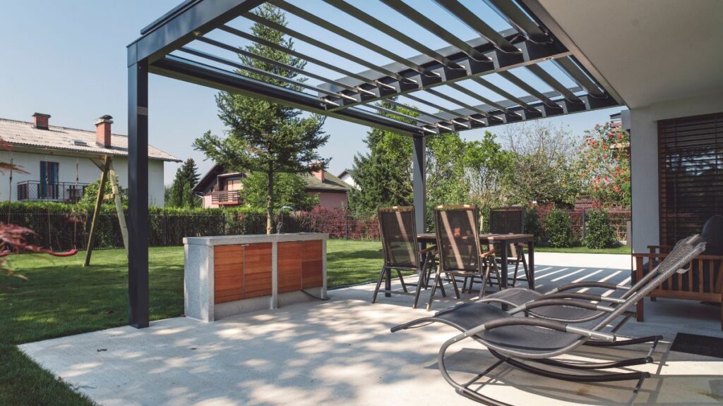 Modern pergola egy tágas teraszon, körülötte zöld növényzet és bútorok.