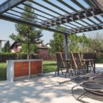 Modern pergola egy tágas teraszon, körülötte zöld növényzet és bútorok.